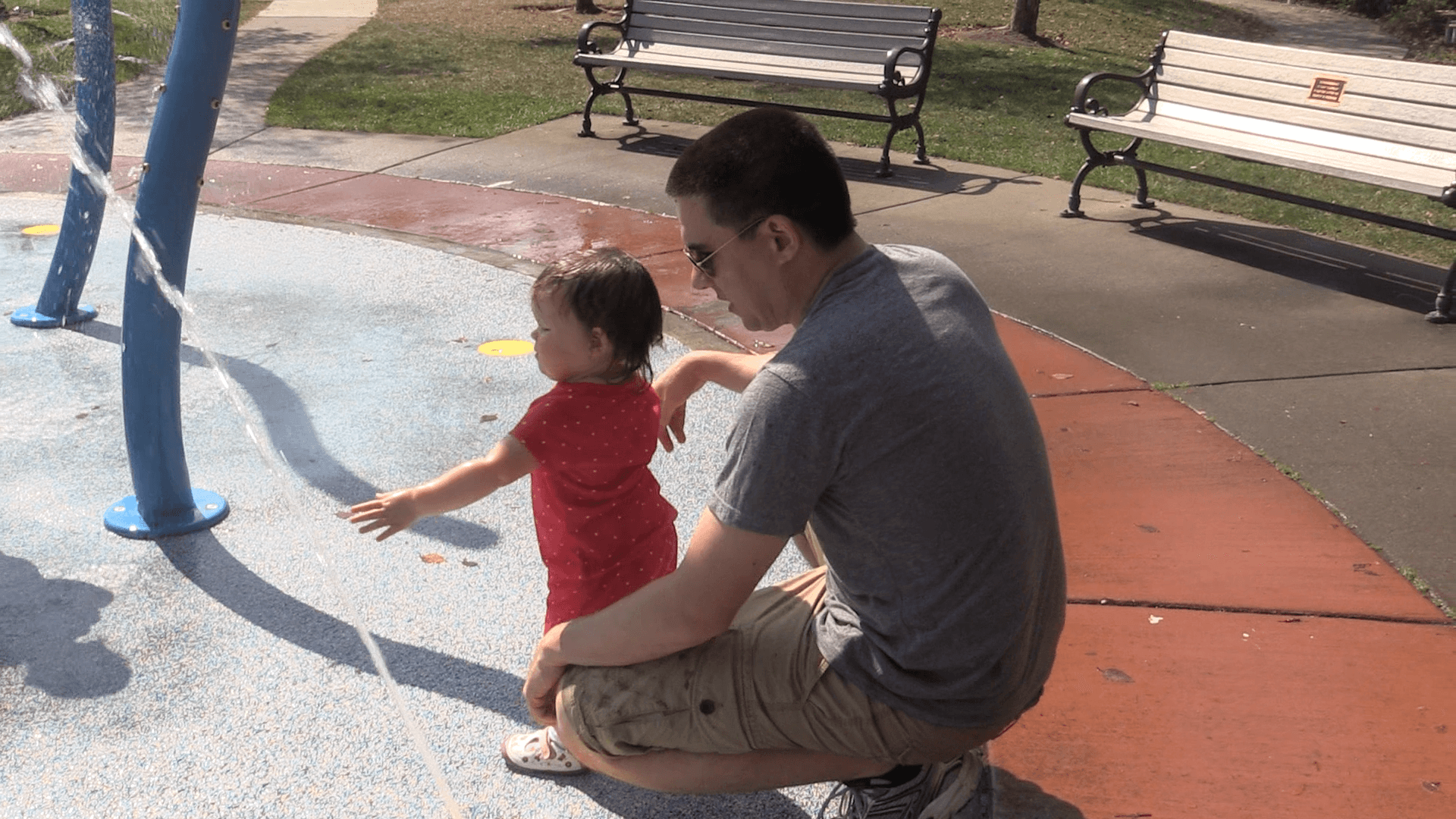 A toddler girl and her dad play at a splash pad in a park.