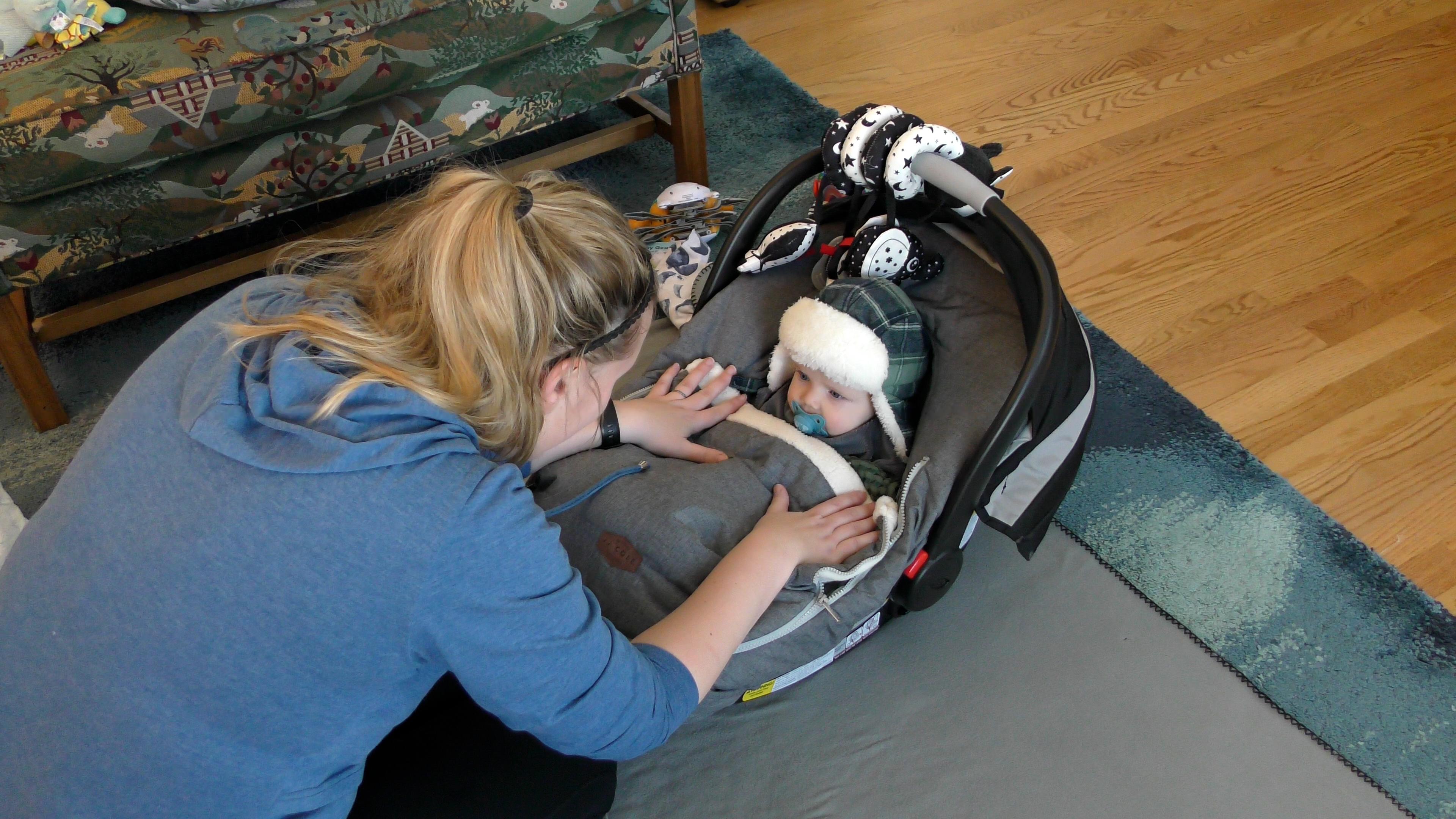 Mom putting a blanket over her baby boy while he is in his car seat on the living room floor. The baby is wearing a green plaid winter hat and sucking on a blue pacifier.